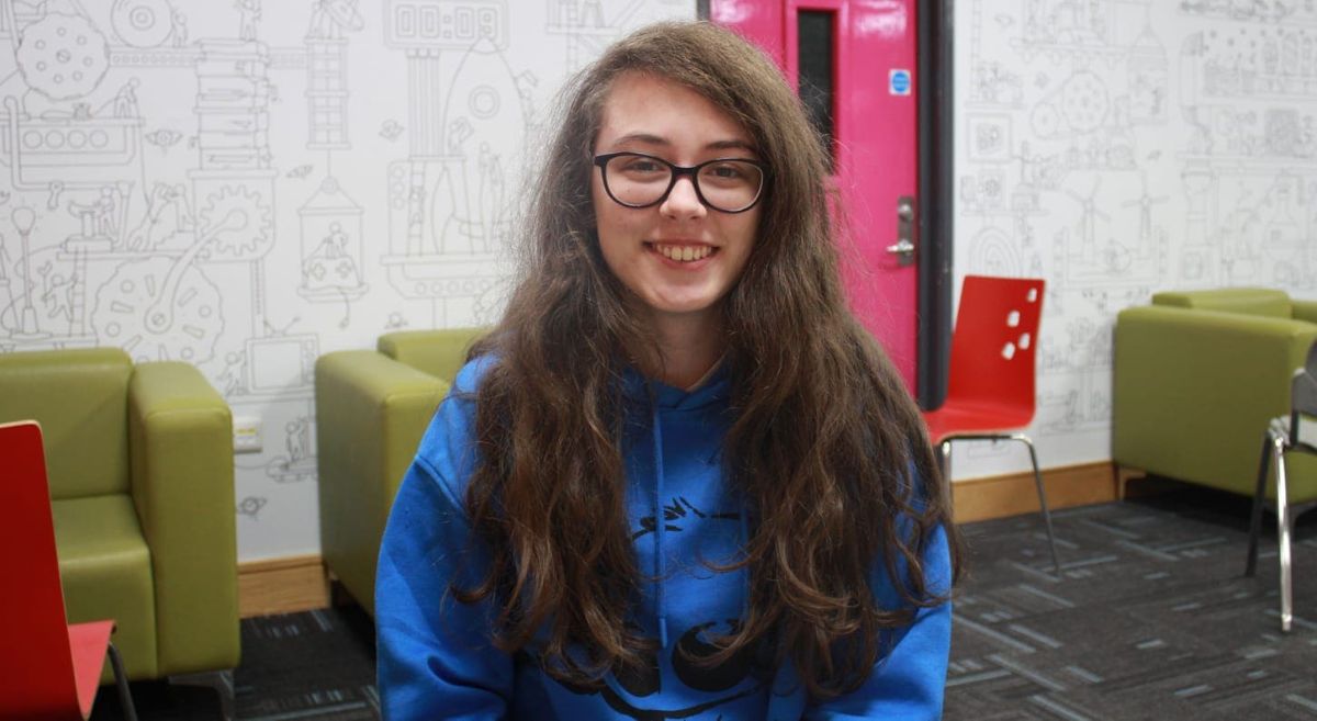 Female student with dark hair, wearing glasses and a blue hoodie, with chairs in the background.